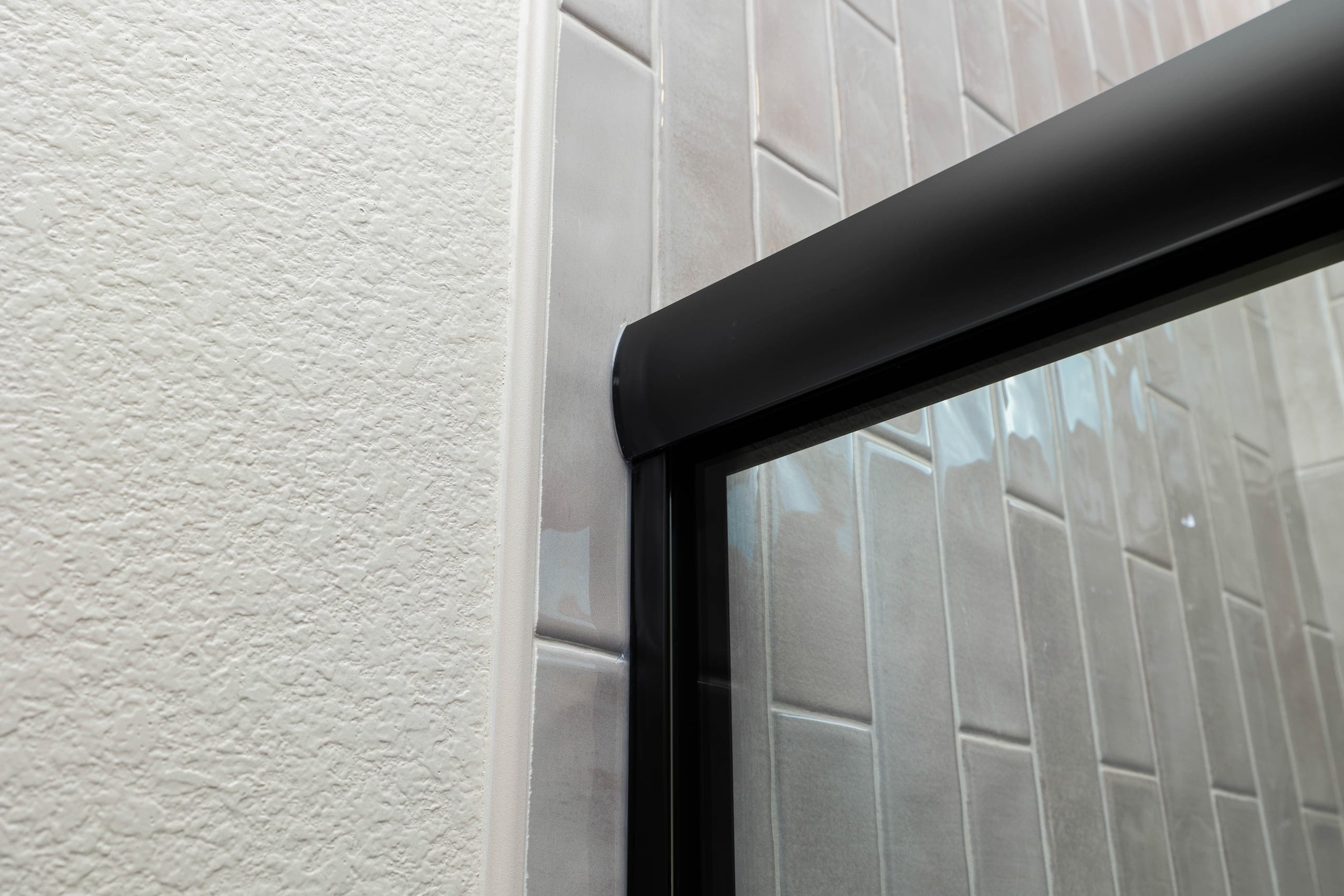Top corner detail of a black-trimmed frameless shower against grey subway tile